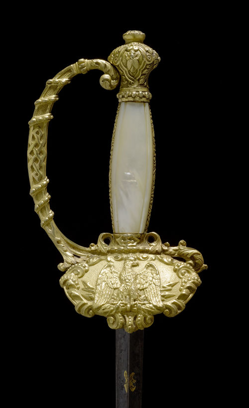 SWORD,France, Napoleon III. Gilt brass. Handle decorated with 2 mother-of-pearl plates. Urn-shaped pommel. Knuckle guard transitioning into cross guard.  Guard plate with eagle. Triangular blade, decorated with trophies (L 80 cm). Black leather scabbard. L in total 98.5 cm.